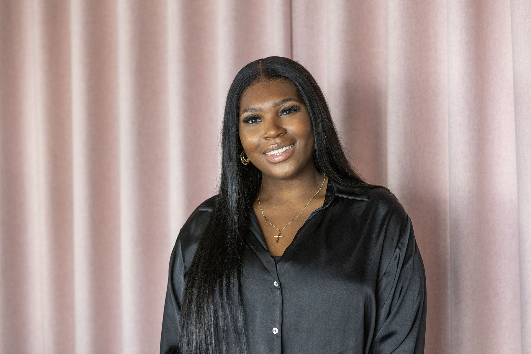 Professional headshot of a smiling woman with long dark hair wearing a black satin blouse and gold jewellery, standing in front of soft blush pink curtains.