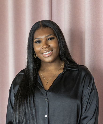 Professional headshot of a smiling woman with long dark hair wearing a black satin blouse and gold jewellery, standing in front of soft blush pink curtains.