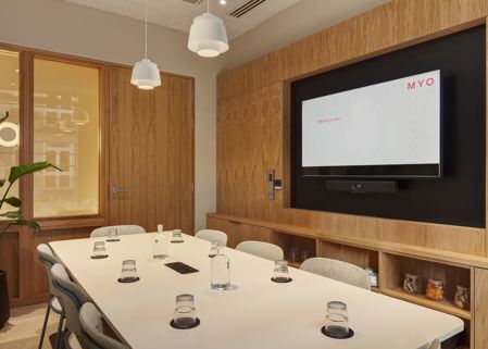 A contemporary meeting room featuring a conference table, upholstered chairs, a wall-mounted presentation screen, timber wall panelling and pendant lighting.