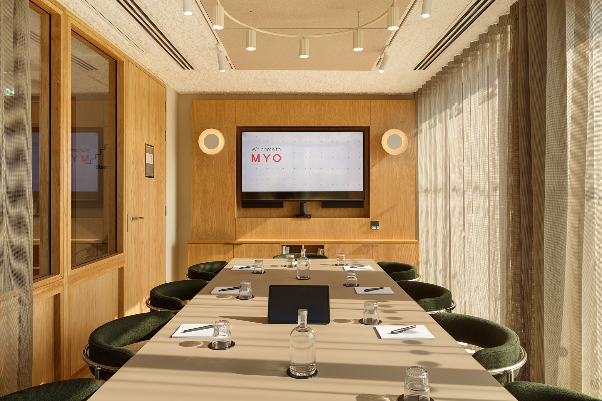 A contemporary boardroom featuring a conference table, green upholstered chairs, a presentation screen, timber wall panelling and floor-to-ceiling windows with sheer curtains.