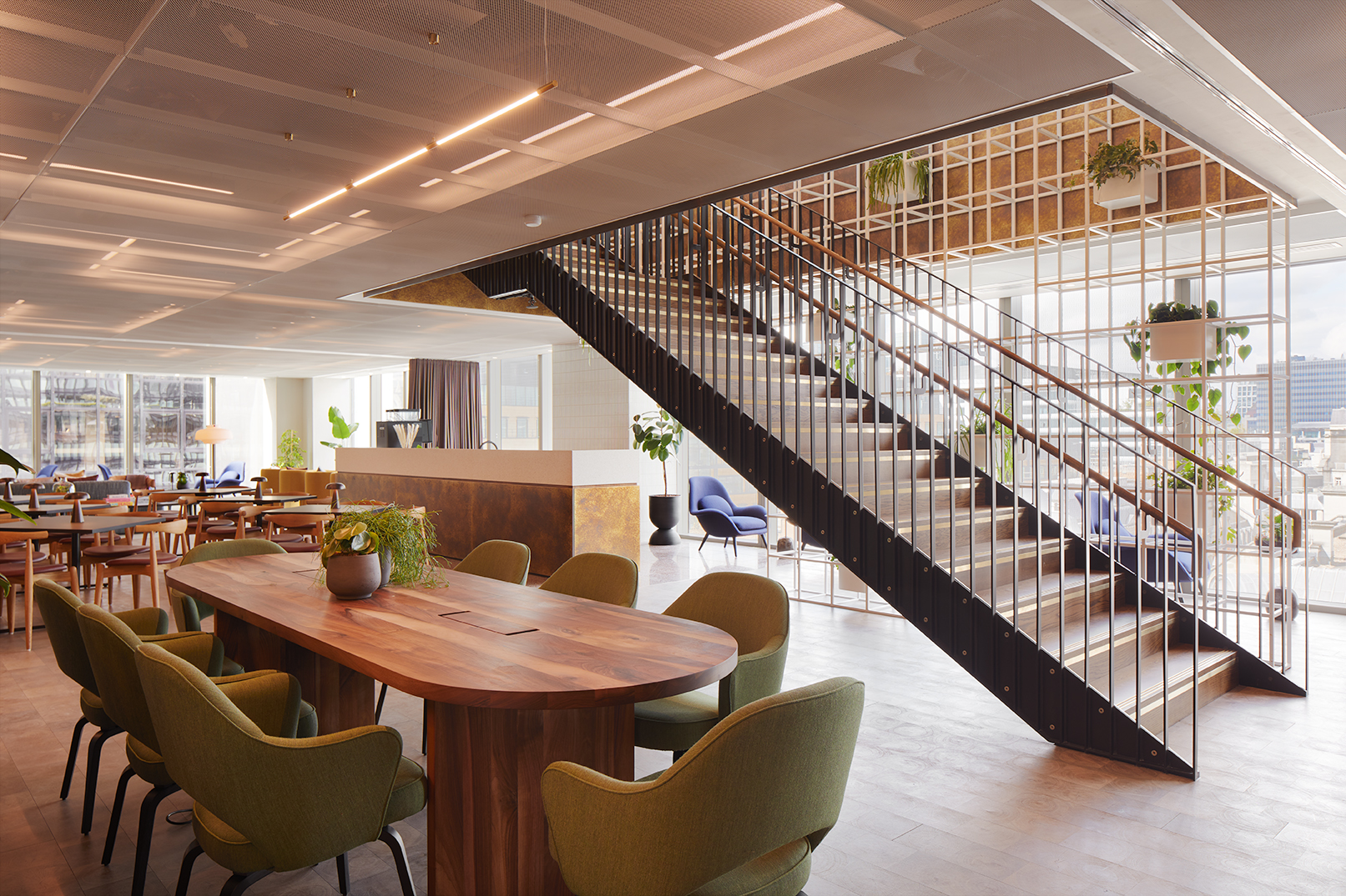 Open lounge area at MYO Liverpool Street featuring a large walnut meeting table with olive green chairs, a statement staircase with black metal detailing, lounge seating and abundant natural light.