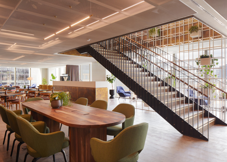 Open lounge area at MYO Liverpool Street featuring a large walnut meeting table with olive green chairs, a statement staircase with black metal detailing, lounge seating and abundant natural light.