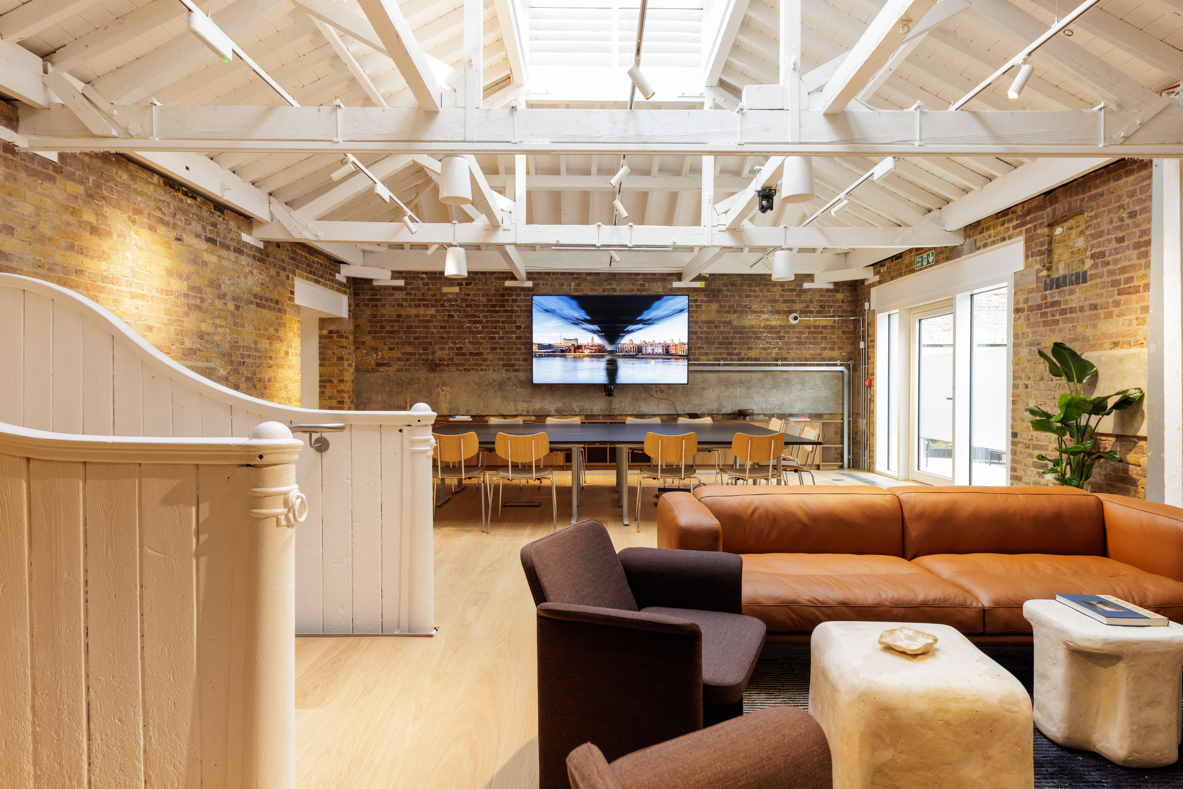 Open lounge workspace with leather sofa, armchairs, dining-style tables and chairs, exposed brick walls and a vaulted white ceiling.