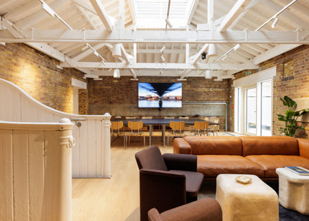 Open lounge workspace with leather sofa, armchairs, dining-style tables and chairs, exposed brick walls and a vaulted white ceiling.
