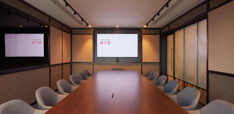 Boardroom at MYO Liverpool Street featuring a large walnut conference table, soft grey upholstered chairs, integrated presentation screen and warm ambient lighting.