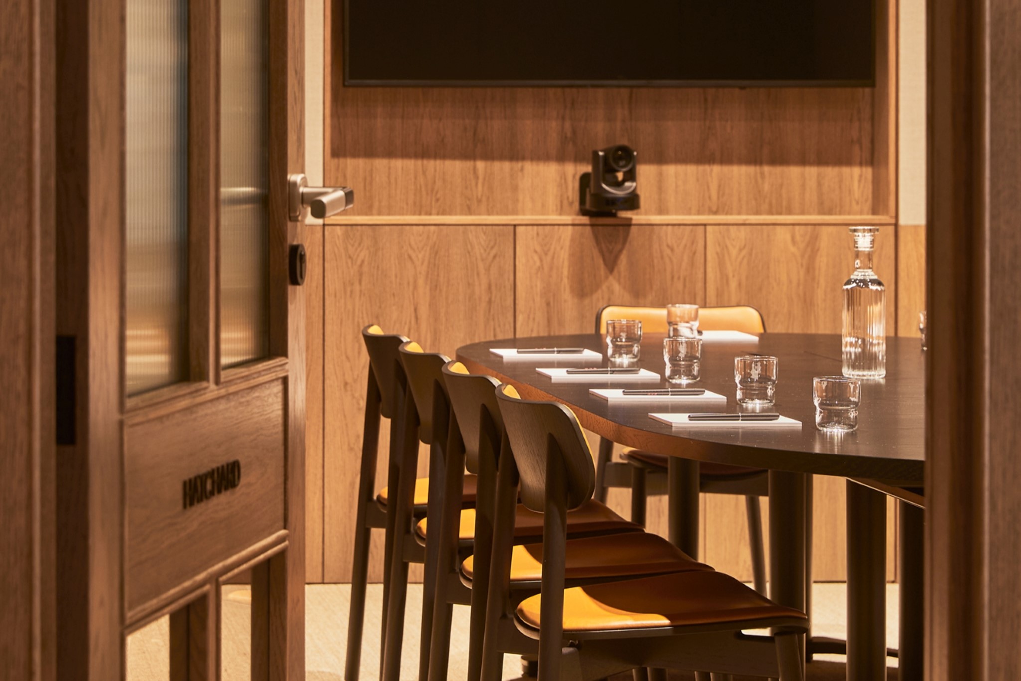 A contemporary meeting room viewed through an open doorway, featuring a round conference table, wooden chairs, video conferencing equipment and a wall-mounted presentation screen.