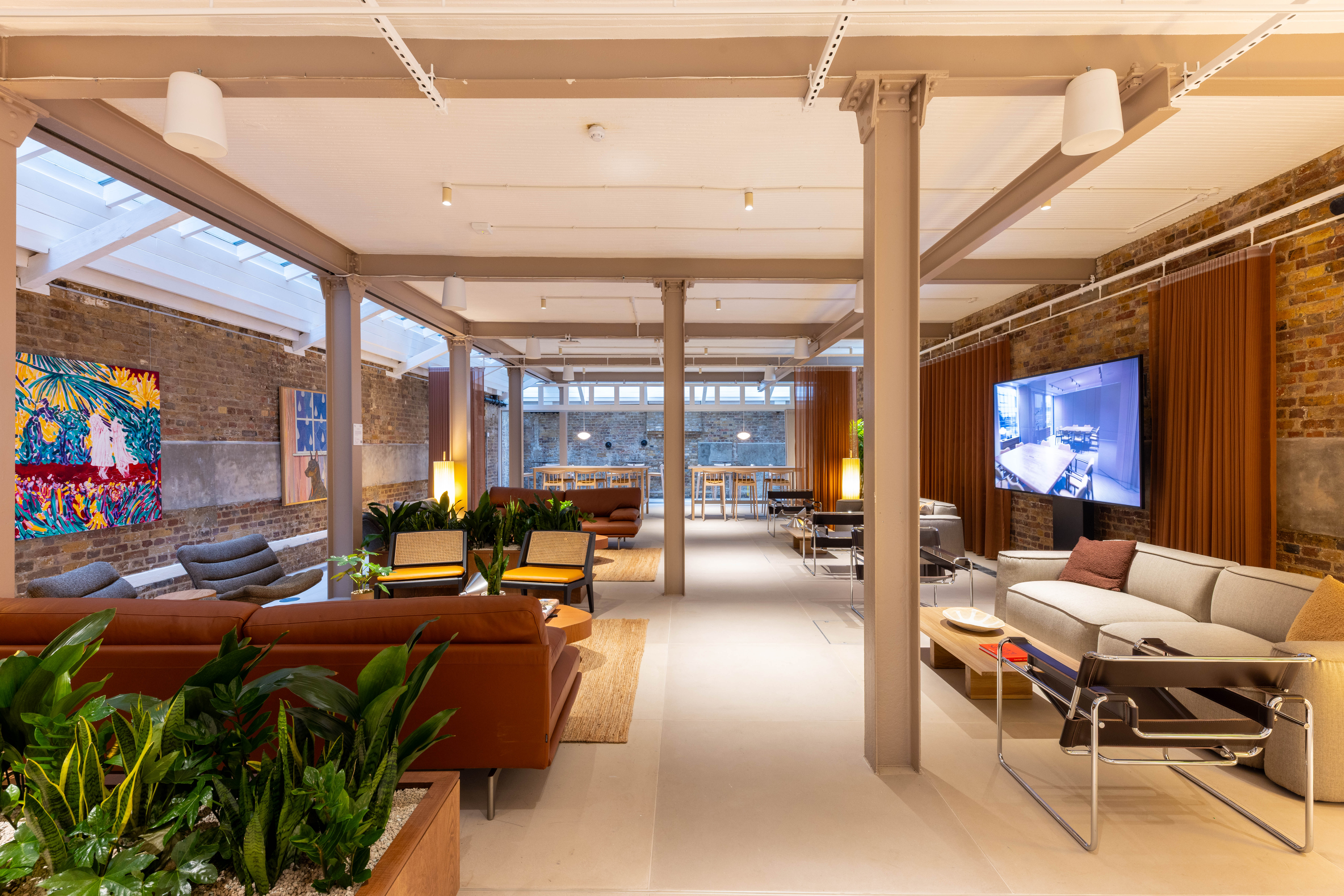 A contemporary communal lounge with sofas, armchairs, indoor planting, exposed brick walls, artwork and a large display screen in a modern workspace.