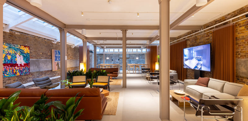 A contemporary communal lounge with sofas, armchairs, indoor planting, exposed brick walls, artwork and a large display screen in a modern workspace.