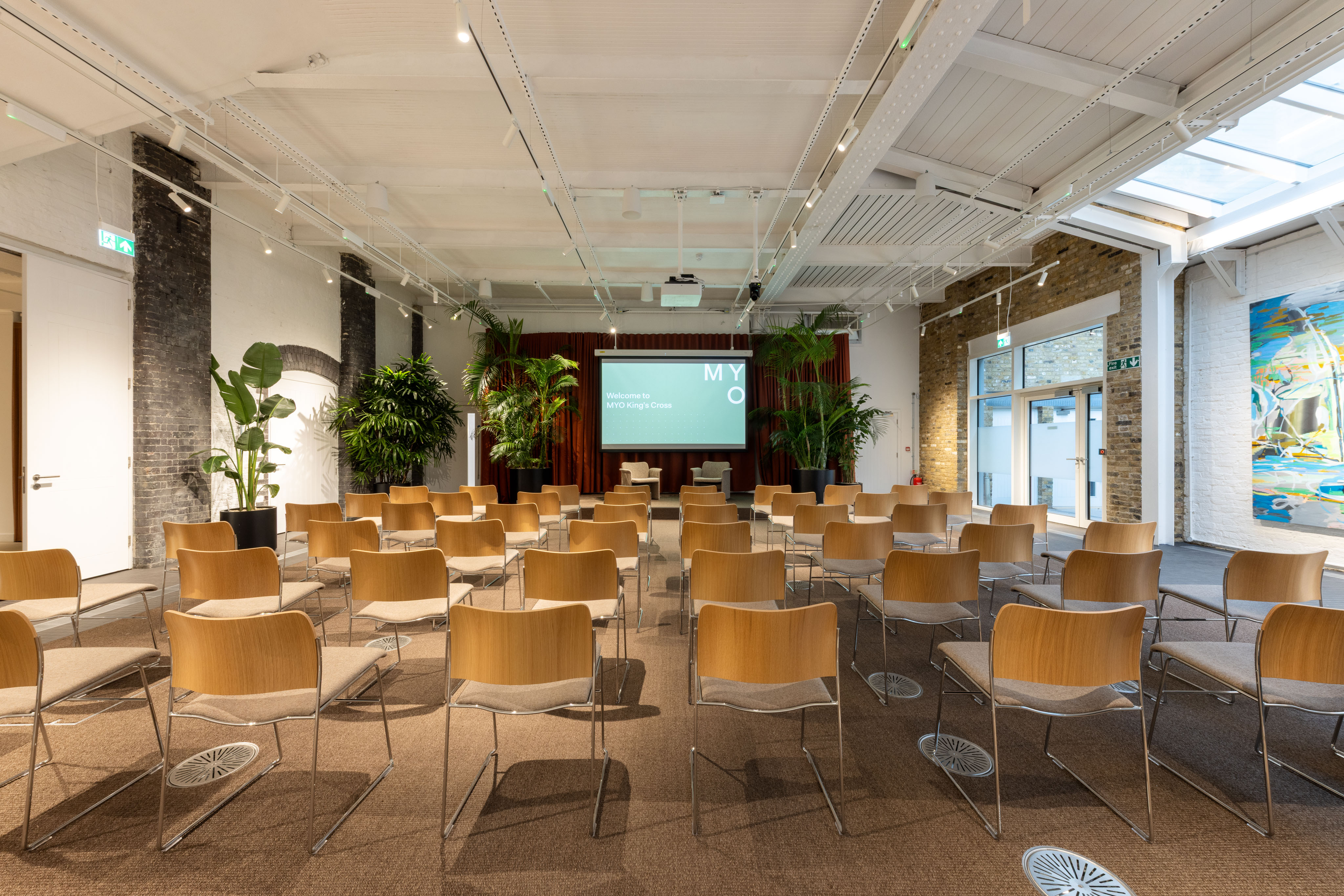 Event room with rows of wooden chairs facing a presentation screen, surrounded by indoor plants, exposed brick walls and a white ceiling with skylights.
