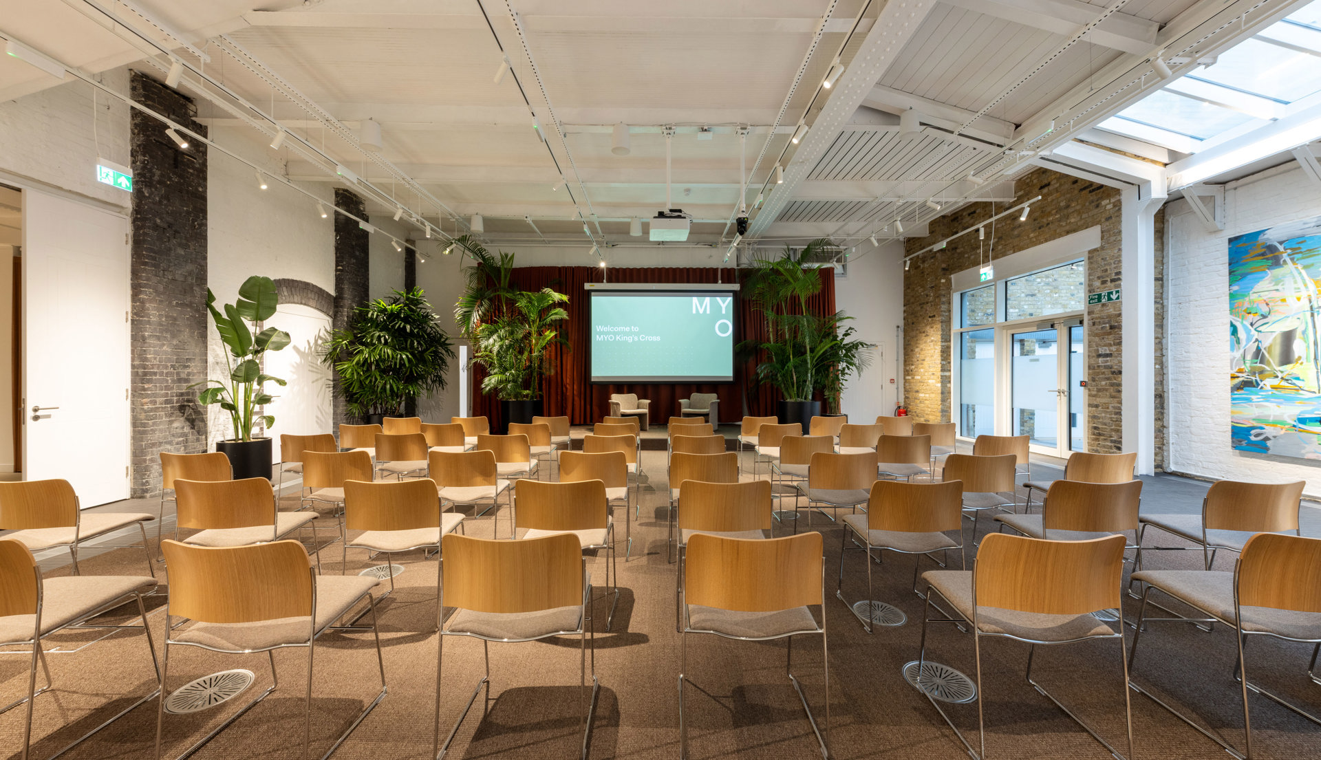 Event room with rows of wooden chairs facing a presentation screen, surrounded by indoor plants, exposed brick walls and a white ceiling with skylights.